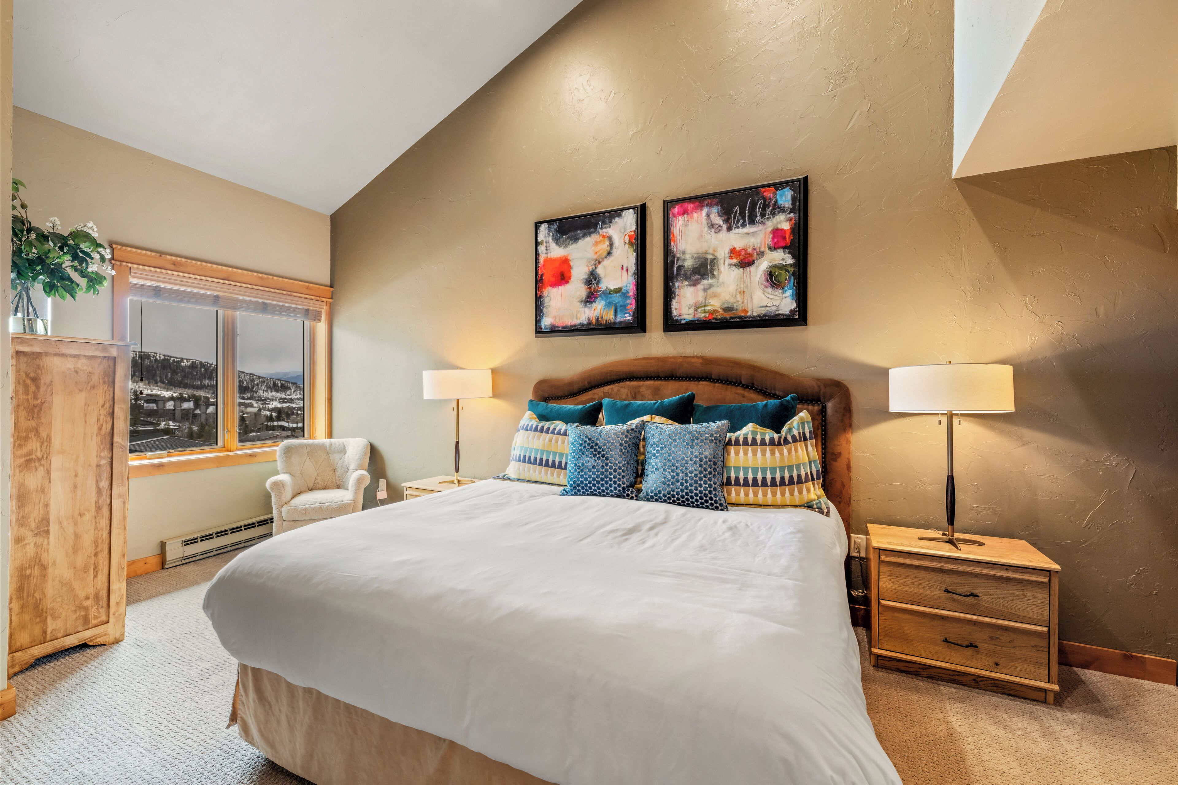 A bedroom with a large bed, two bedside lamps, a wooden nightstand, a chair, a window with a view, and two abstract paintings on the wall.