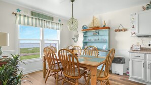 Un comedor con mesa y sillas de madera, vista al exterior, un armario azul con adornos náuticos y un cartel que dice 'Welcome' sobre la ventana.