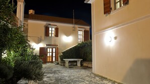 A stone-paved courtyard with a bench, a small table, and a building with a tiled roof and wooden shutters.