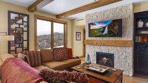 Un salón con chimenea de piedra, un televisor grande montado en la pared, un sofá con cojines estampados y vistas a la ciudad a través de un ventanal.