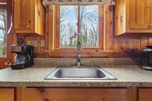 A kitchen with wooden cabinets, a granite countertop, a stainless steel sink, a coffee maker, and a window with a view of trees.