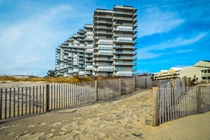 A modern high-rise building with multiple balconies, situated on a sandy beach with a wooden fence in the foreground. - Colorful, spacious 2 bedroom oceanfront condo with WiFi, an outdoor pool, and a great ocean view located midtown and just steps to the beach! (Ocean City)