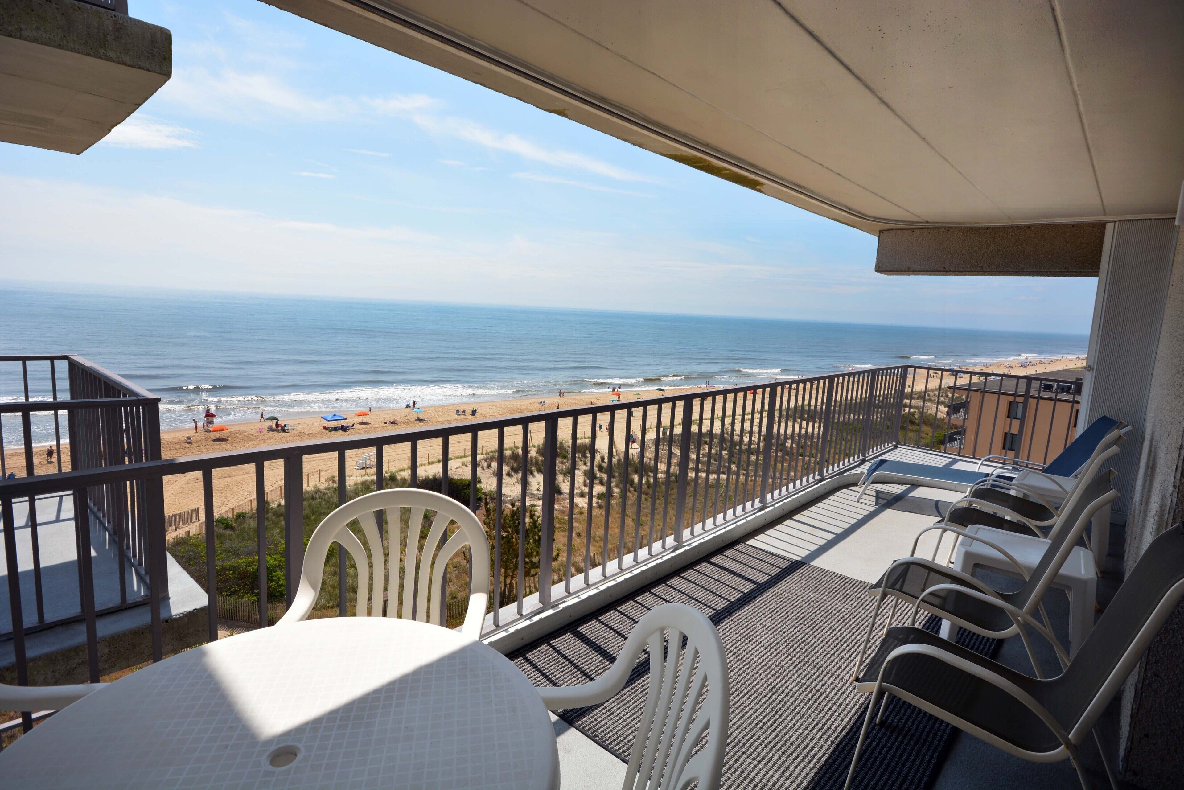 A balcony with a view of the beach and ocean. There are white chairs and a table on the balcony.