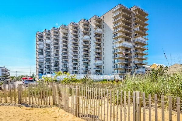 A high-rise residential building with multiple balconies, a sandy beach in the foreground, and a clear blue sky.