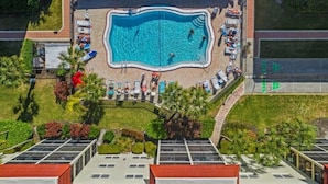 Luftaufnahme eines Schwimmbeckens, umgeben von Liegestühlen und Sonnenschirmen, mit blauem Himmel.