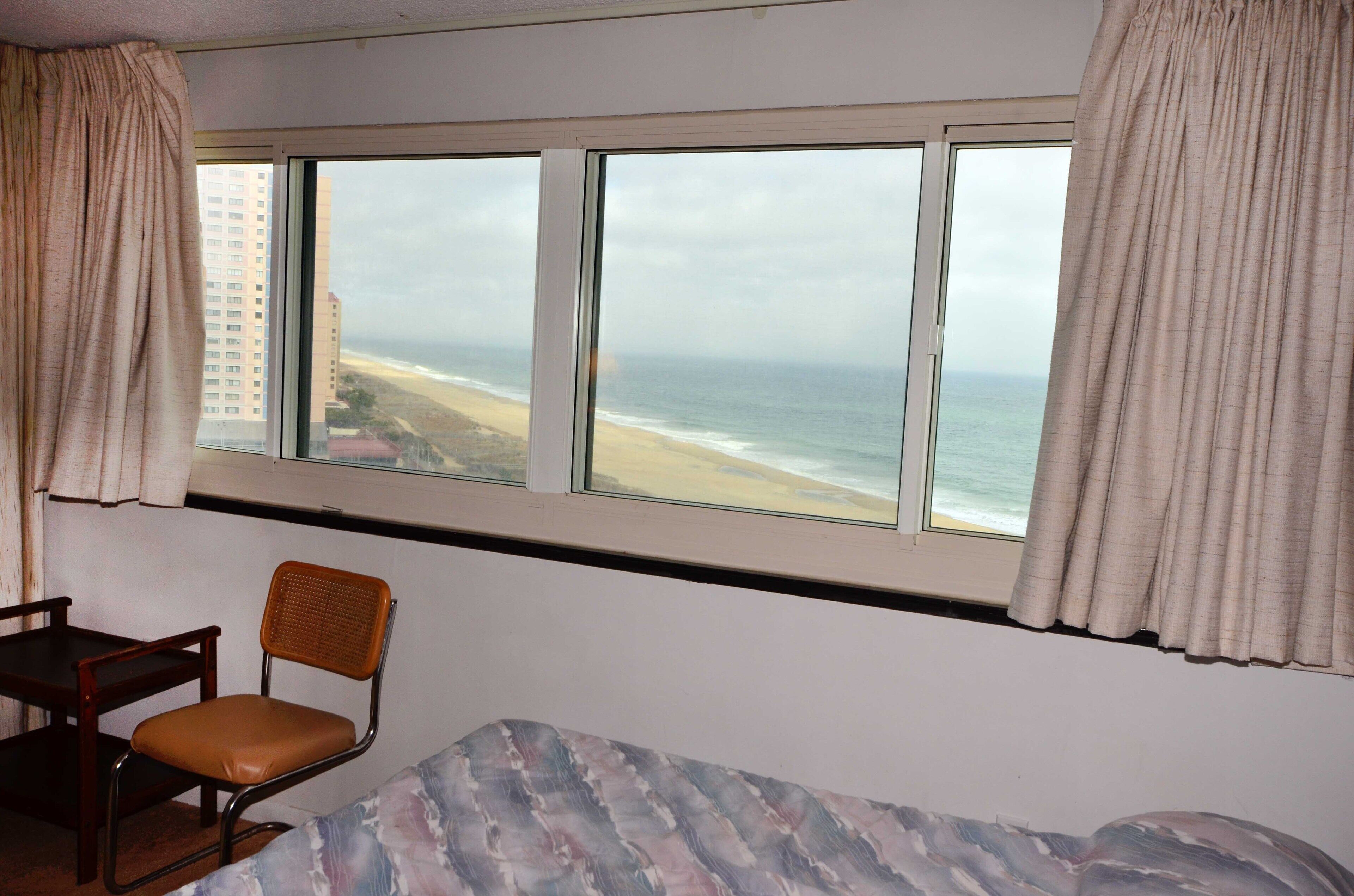 A room with a large window overlooking a beach, a chair, and a bed.