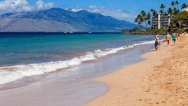 A beach with clear blue water, a mountain in the background, and a few people walking along the shore. - Tranquil Courtyard View Condo w/Kitchen Updates, WiFi, Washer/Dryer–#7203 (KIHEI)