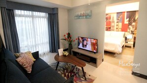 Ein modernes Wohnzimmer mit einer Couch, einem Fernseher und einem kleinen Couchtisch. An der Wand hängt ein Gemälde, und im Hintergrund steht ein Bett.