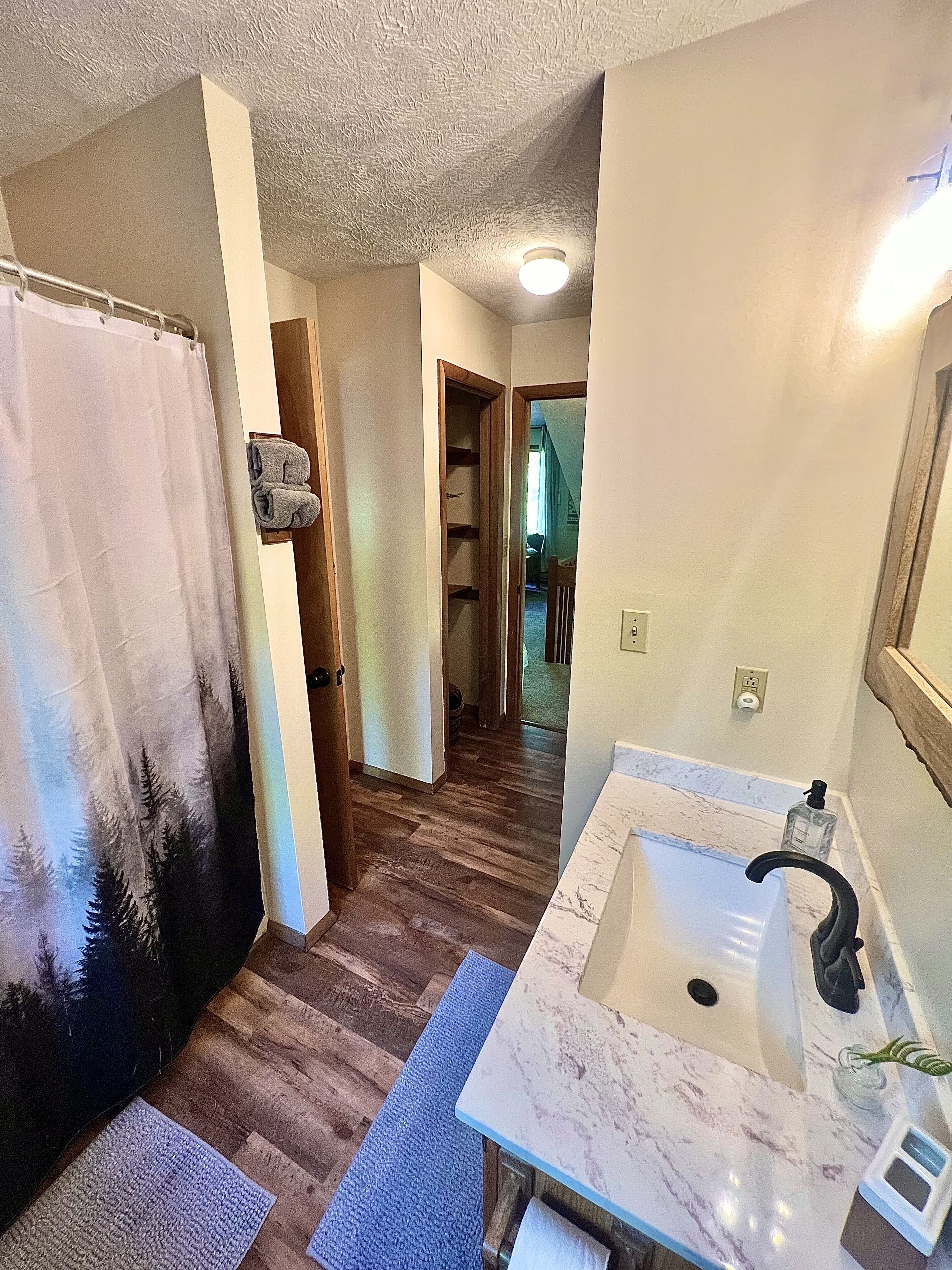 Un baño con lavamanos de mármol, suelo de madera y cortina de ducha con diseño de bosque.