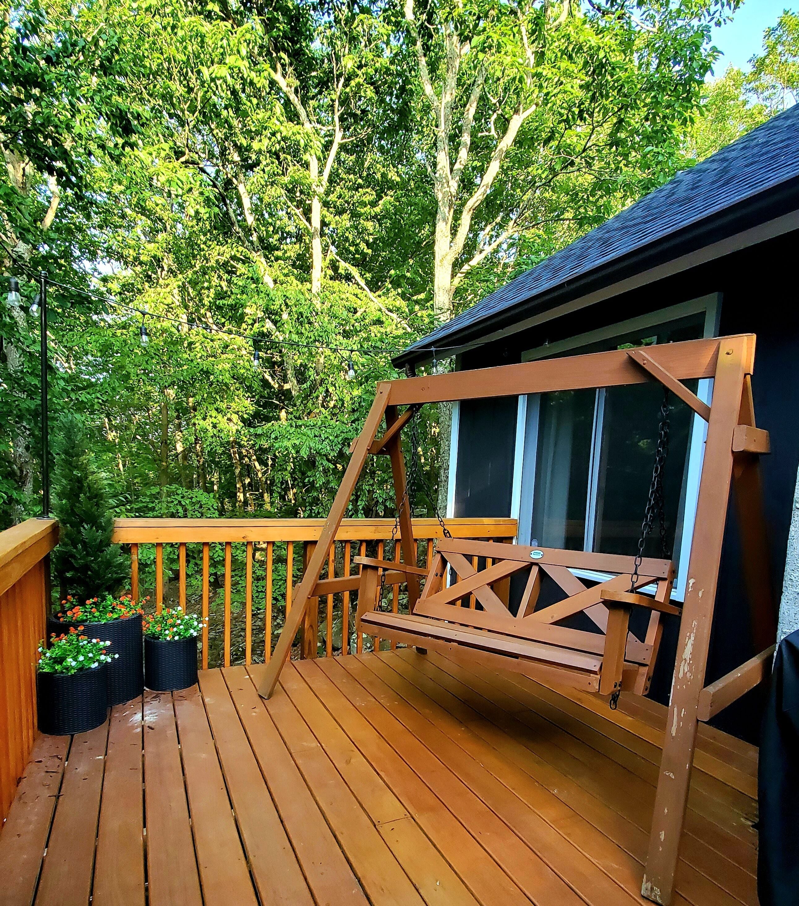 A wooden deck with a swing set, surrounded by lush greenery and a building with large windows.
