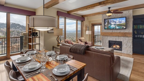 A dining area with a wooden table set for six, a fireplace, and a large TV mounted on a stone wall.