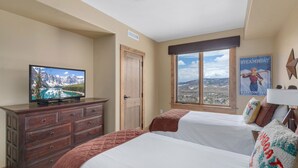 A hotel room with two beds, a wooden dresser, a television displaying a mountain landscape, a window with a view of a cityscape, and a poster on the wall.
