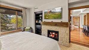 Ein Schlafzimmer mit Kamin aus Stein, einem großen Bett und einem Fernseher, der über dem Kamin angebracht ist, sowie mit Blick ins Freie durch ein Fenster.