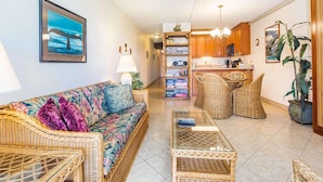 A living room with a sofa, coffee table, and wicker chairs. There is a kitchen area with wooden cabinets and a dining table. - Island Energy in Suite w/Lanai, Washer/Dryer, WiFi, Kitchen–Kamaole Sands 7302 (KIHEI)