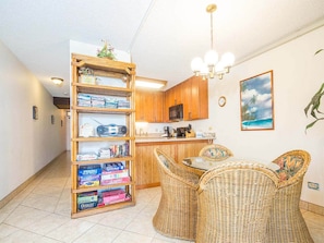A kitchen with wooden cabinets, a dining table with wicker chairs, and a bookshelf filled with board games.