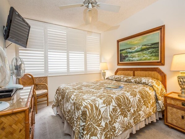 A bedroom with a bed, a television, a fan, a chair, and a painting on the wall. - Island Energy in Suite w/Lanai, Washer/Dryer, WiFi, Kitchen–Kamaole Sands 7302 (KIHEI)