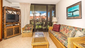 A living room with a television, wicker furniture, a sofa with floral upholstery, and a view of a residential area through a sliding door. - Island Energy in Suite w/Lanai, Washer/Dryer, WiFi, Kitchen–Kamaole Sands 7302 (KIHEI)