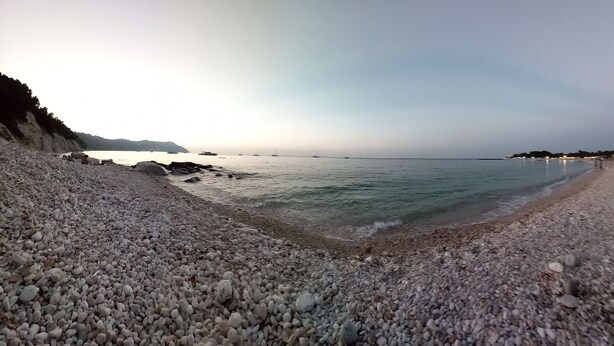 Une plage de galets avec une eau calme et des bateaux qui se dessinent à l’horizon.