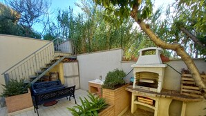 Un jardin avec un espace barbecue en briques, une table en bois avec un banc et un escalier menant à un porche abrité.