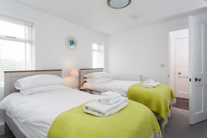 A hotel room with two beds, a bedside table with a lamp, a window with blinds, a door, and a wall-mounted light fixture.