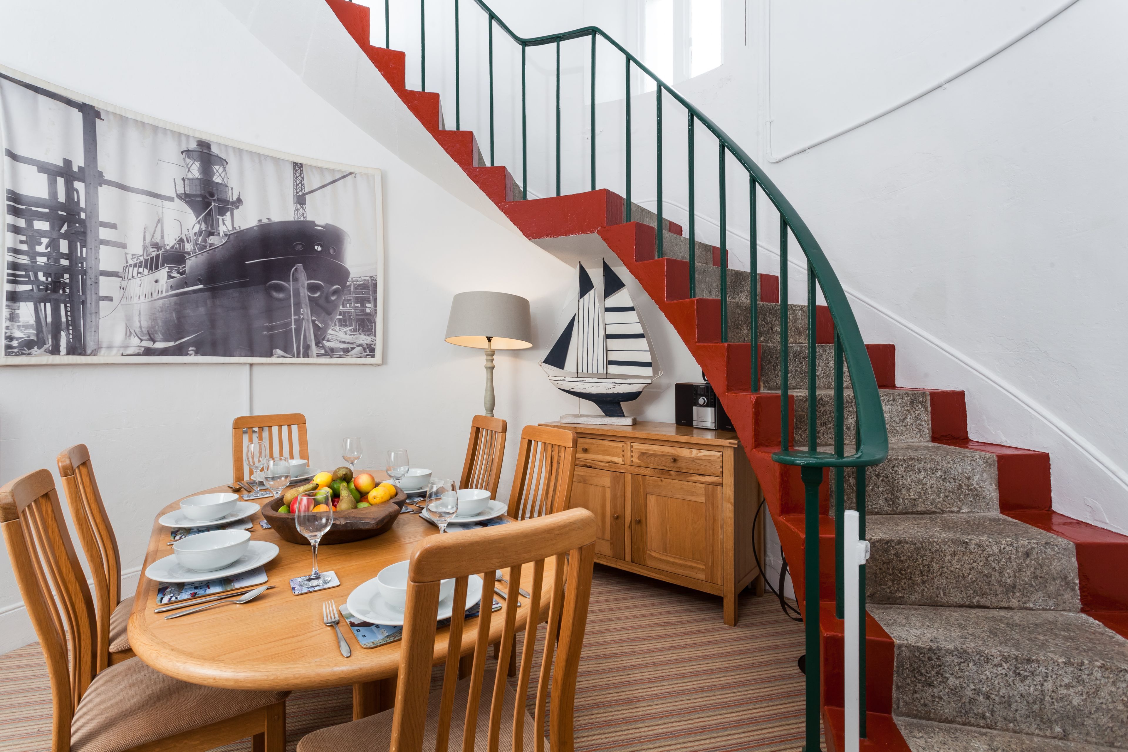 Un comedor con una mesa de madera dispuesta para cuatro personas, rodeada de sillas. Una escalera con barandas rojas y verdes conduce a un nivel superior.
