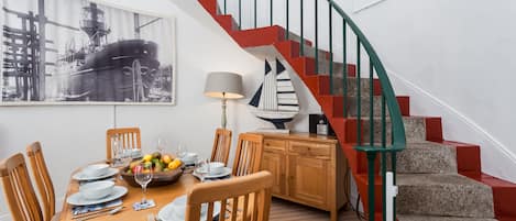 A dining area with a wooden table set for four, surrounded by chairs. A staircase with red and green railings leads to an upper level.