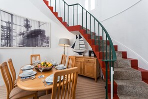 A dining area with a wooden table set for four, surrounded by chairs. A staircase with red and green railings leads to an upper level.