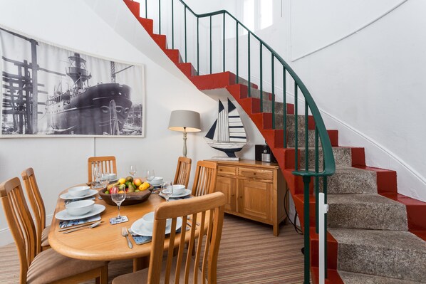 A dining area with a wooden table set for four, surrounded by chairs. A staircase with red and green railings leads to an upper level.