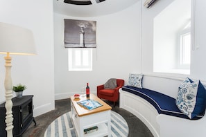 A modern living room with a fireplace, a red armchair, a sofa with nautical-themed cushions, a painting of a lighthouse, and a dining table with a bottle of wine.