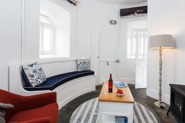 A room with a curved bench, a wooden table with a bottle and glasses, a lamp, and a fireplace.