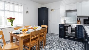 A modern kitchen with a dining area, featuring a black cabinet, white cabinets, a patterned floor, and a window with blinds.
