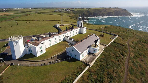 A coastal building with a lighthouse, surrounded by green fields and the ocean.