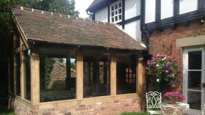 A conservatory with a brick base and wooden framing, a small garden area with a table and chairs, and a flower basket on the right. - Luxury Coach House in beautiful rural village, scenic walks & 3 miles to Ledbury (Ledbury)