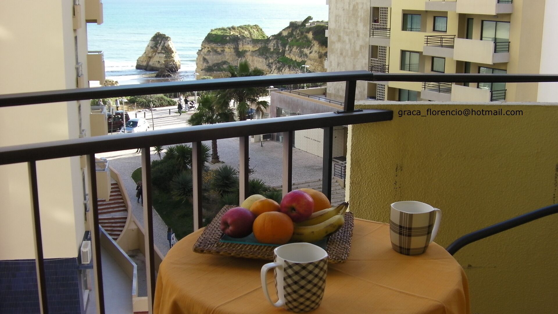 A balcony with a table set with fruits and two mugs, overlooking a coastal area with buildings and cliffs.