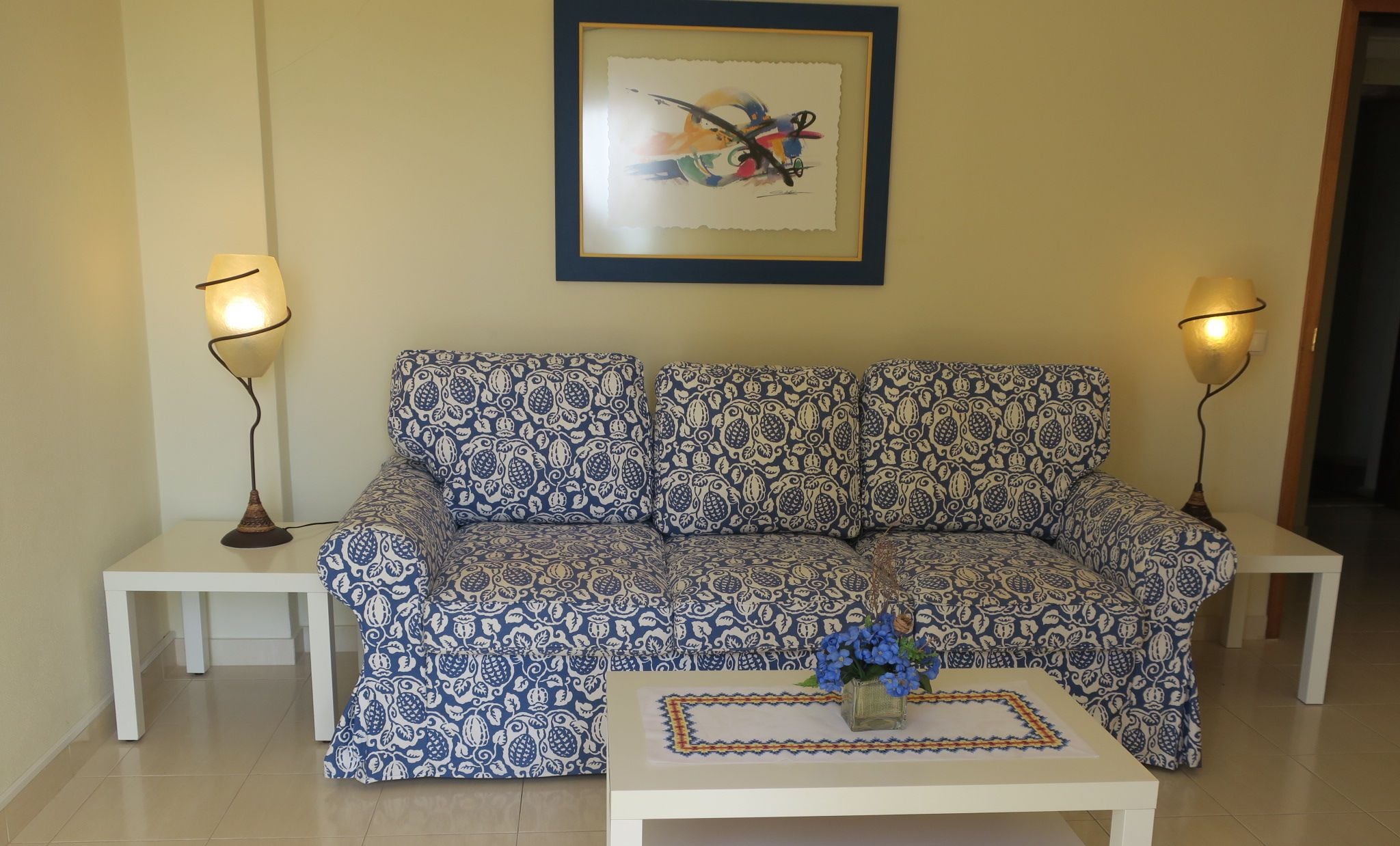 A living room with a patterned sofa, two side tables, a coffee table with a vase, and two floor lamps.