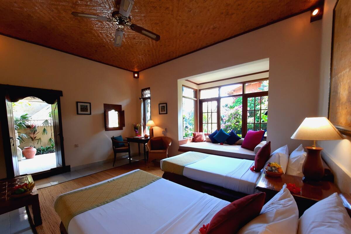 A hotel room with two beds, a ceiling fan, a window with a view of greenery, and a table with a lamp.