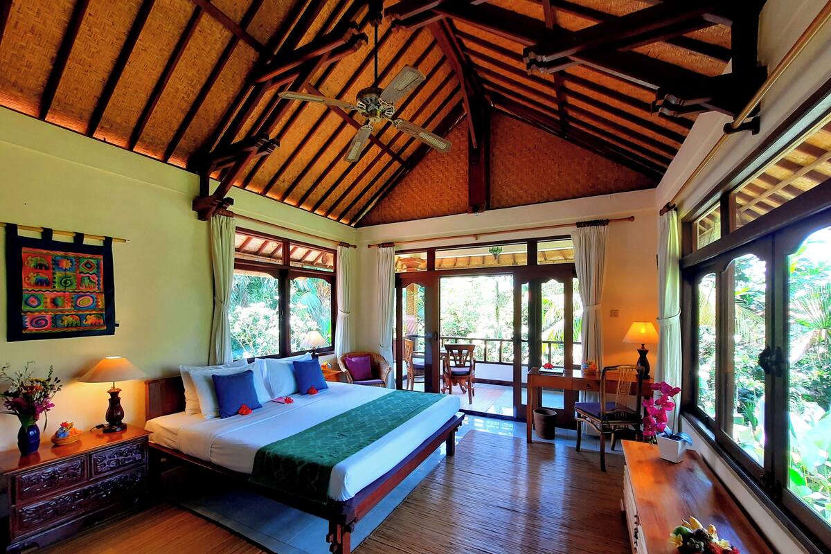 A spacious bedroom with a large bed, wooden ceiling beams, and a view of lush greenery outside.