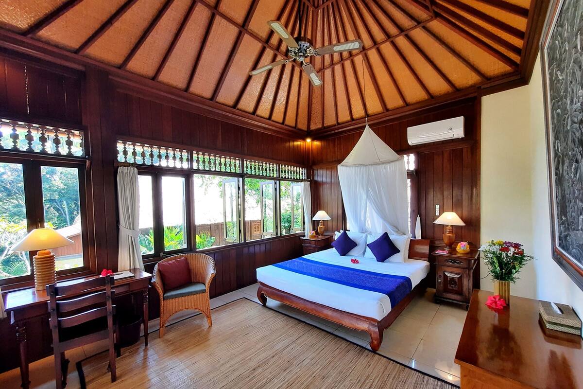 A bedroom with a wooden ceiling, a bed with a canopy, two bedside tables with lamps, a chair, and a desk.
