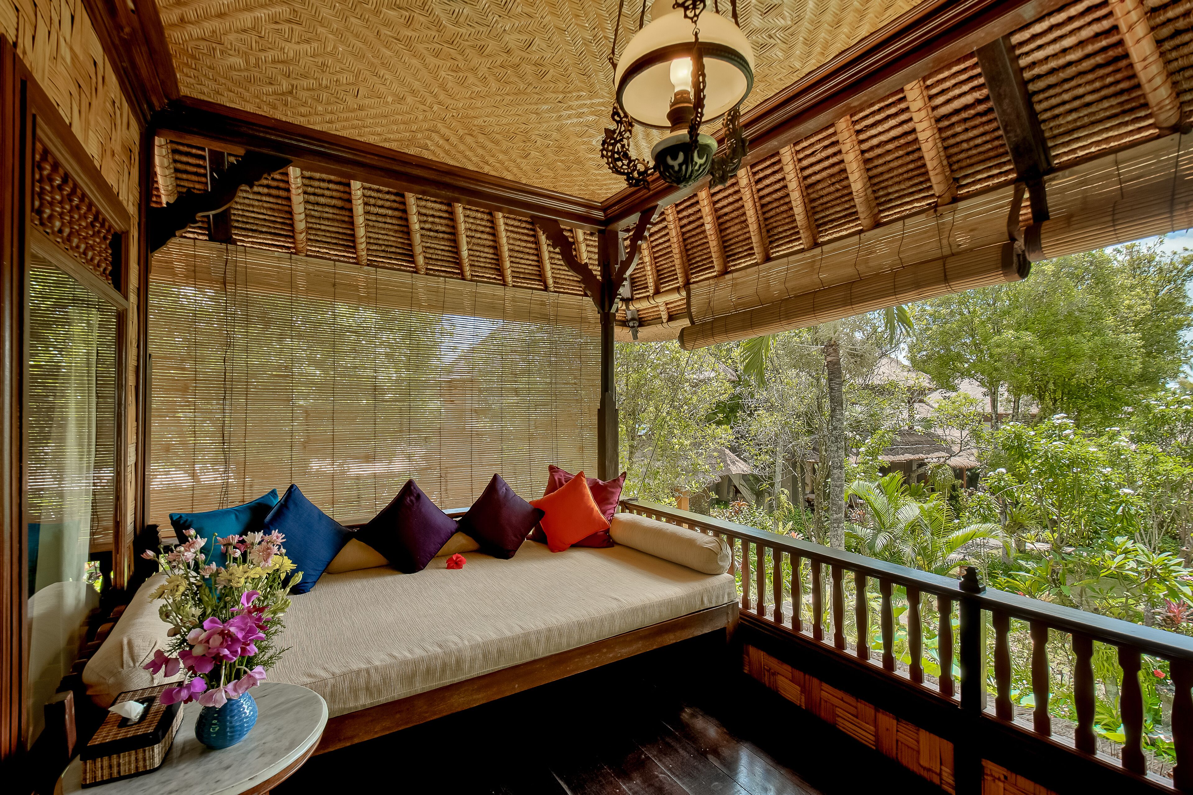 Una habitación con techo de madera, un banco acolchado con almohadas, una mesita con un jarrón y vistas al exterior con vegetación.