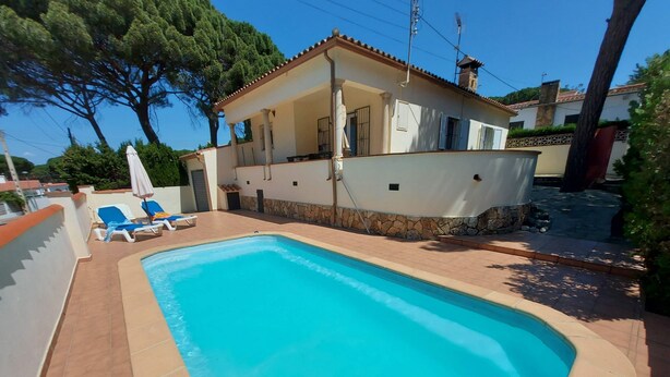 A rectangular swimming pool with a tiled deck, surrounded by a white building with a tiled roof and stone accents. There are two lounge chairs with blue cushions and a white umbrella.