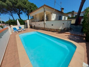 Ein rechteckiger Pool mit gefliestem Sonnendeck, umgeben von einem weißen Gebäude mit einem gefliesten Dach und Steindekorationen. Zwei Liegestühle mit blauen Kissen und einem weißen Sonnenschirm sind vorhanden.