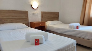 A hotel room with two beds, each with neatly made white linens and towels. There is a wooden nightstand between the beds, and a wall-mounted light fixture above the nightstand.