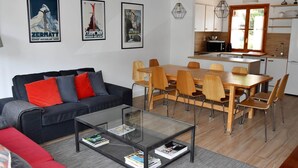 Een moderne woonkamer met een zwarte bank, rode en grijze kussens, een glazen salontafel en een houten eettafel met zes stoelen. Op de muur hangen ingekaderde posters, er is een keuken met witte kasten en een raam met uitzicht op groen.