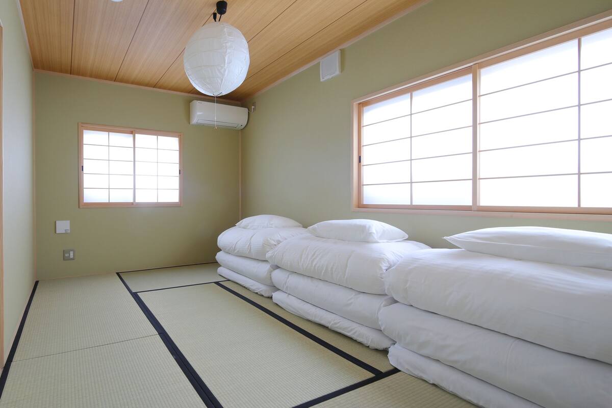 Una habitación con suelo de tatami, un ventanal con biombos y una pila de ropa de cama blanca.