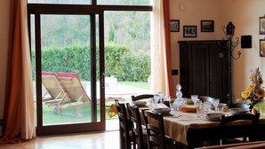 A dining area with a table set for four, a view of the garden through a sliding glass door, and a dark wooden cabinet against the wall. - Villa Gilda Bracciano with private Pool & A/C (Bracciano, Italia)