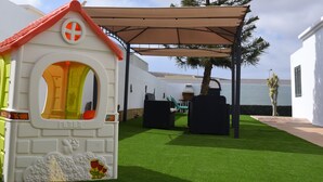 A children's playhouse with a red roof and a yellow door, set on artificial grass with a shaded area nearby. - Villa Nirvan with Heated Pool (playa blanca, yaiza, lanzarote)