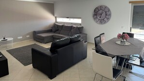 A modern living room with a grey sectional sofa, a dining table with chairs, a round wall clock, and a window with blinds. - Modern apartment 5 km from Carvoeiro with terrace and barbecue (Lagoa)