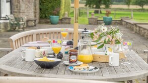 Et bord dækket udendørs med morgenmad, herunder en skål chips, et glas appelsinjuice, en flaske marmelade og en kande vand.
