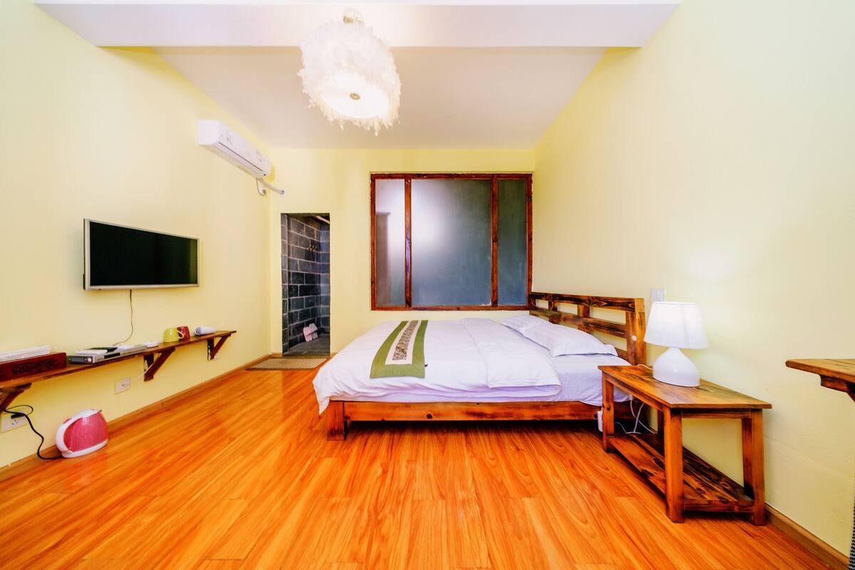 A bedroom with a wooden bed, a bedside table with a lamp, a TV mounted on the wall, and a shelf with various items.