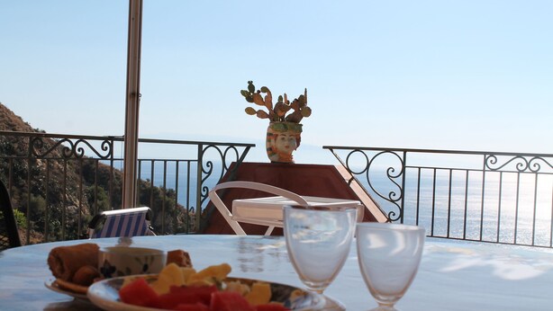 A table with a bowl of fruit, a glass, and a cup, set on a balcony overlooking a coastal view.